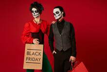 Stylish Couple In Dia De Los Muertos Skull Makeup Standing With Shopping Bags On Red, Black Friday