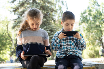 Wall Mural - Children use gadgets while sitting in the park outdoors, games on smartphone and tablet