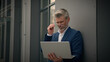 © Yuliia - Caucasian mature retired business man with laptop working distant online work in city leaning to wall off company office building manager senior businessman with computer outdoors typing free wi-fi