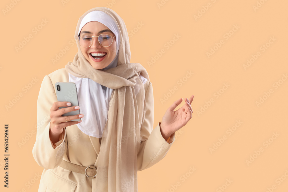Young Muslim woman in stylish suit using mobile phone on beige background