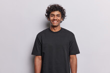 People Positive Emotions Concept. Studio Waist Up Of Young Happy Smiling Broadly Hindu Man Standing In Centre Isolated On White Background Wearing Black Casual T Shirt Looking Straight At Camera