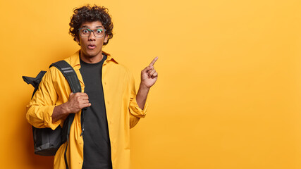 Wall Mural - People emotions concept, Studio shot of young surprised Hindu male student standing isolated on left on yellow background pointing at blank space for your promotion wearing casual clothes with bag