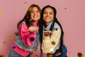 Poster - Two joyful young women blowing colorful confetti and smiling against pink background