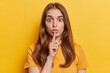 Studio close up of young beautiful European lady with long loose brunette hair in yellow t shirt standing in centre isolated making gesture to keep silence and be quiet and still looking straight