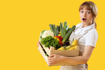 Wall Mural - Shocked young female farmer with wooden box full of different ripe vegetables on yellow background