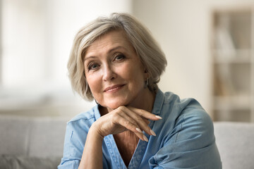 Positive pretty elder senior woman looking at camera, leaning chin on hand, wrist in happy thoughts, posing for home portrait, enjoying retirement, promoting modern elderly beauty care