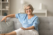 Happy pretty mature old woman in glasses resting on soft couch, looking at camera with toothy smile, looking at camera, posing for portrait, enjoying good retirement, comfortable home