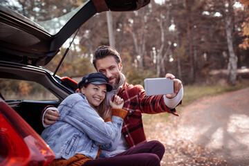 Young Caucasian couple taking a selfie while sitting on the trunk of their car in a forest park