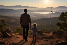 dad holding hands with his son on the top of a mountain looking at the windmills, unrecognizable people, at sunset, wind energy concept