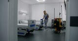 Fototapeta  - Adult nurse mops floor between beds in hospital ward. Health worker brings cleaning trolley to hospital room. Female cleaner prepares ward for new patients. Medical staff at work in modern clinic.