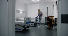 Adult Nurse Mops Floor Between Beds In Hospital Ward. Health Worker Brings Cleaning Trolley To Hospital Room. Female Cleaner Prepares Ward For New Patients. Medical Staff At Work In Modern Clinic.