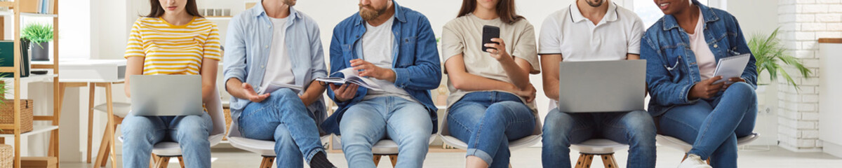 Multiracial people in jeans sitting in row on office chairs, waiting for work meeting, talking and using mobile phones and laptop computers. Crop low section shot, banner. Modern communication concept