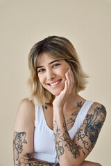 Wall Mural - Happy blond pretty happy girl beauty female model with short blonde hair and tattoos looking at camera touching face pampering advertising skin care dermatology isolated at beige background. Portrait.