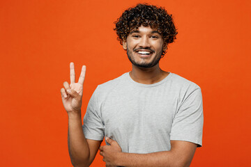 Wall Mural - Young smiling cheerful fun cool joyful happy Indian man he wears t-shirt casual clothes showing victory sign looking camera isolated on orange red color background studio portrait. Lifestyle concept.