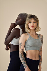 Wall Mural - Two African and Caucsian gen z girls hipster inclusive multiethnic models, cool diverse fit young women wearing sportswear grey tops and leggings standing on beige background, vertical portrait.