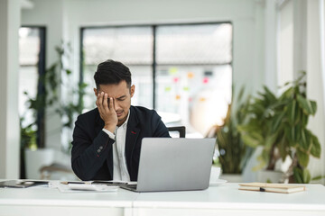 Wall Mural - Young unhappy man office worker feeling bored at work, looking at laptop with demotivated face expression while sitting at workplace in office, distracted male worker feeling tired of monotonous job