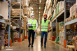 © alfa27 - Retail Warehouse full of Shelves with Goods, Two Caucasian Male Workers Supervisors in Working Uniform and Helmet Discuss Product Delivery. Workers Walking and Having Talk In Warehouse