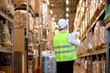 © alfa27 - rear view on young male in helmet with clipboard checking goods at warehouse, counting and controlling distribution. unrecognizable hardworking caucasian worker in green working uniform