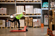 © alfa27 - joyous caucasian male staff dressed in working clothes and helmet in warehouse having fun standing on pallet truck jack, driving among shelves, enjoy free time, leisure at work place