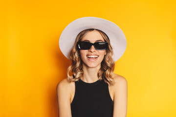 Wall Mural - Close-up portrait of blonde woman casual portrait in positive view, big smile, beautiful model posing in studio over yellow background. Caucasian portrait woman in white hat and black top, eyeglasses.