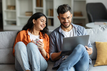 Young hindu couple resting on sofa with laptop, woman drinking coffee, spouses resting in living room, browsing internet together, choosing movie for evening