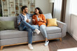 © Home-stock - Happy indian couple relaxing and drinking coffee, sitting on couch at home, talking while resting with hot drinks in cups at modern living room, free space