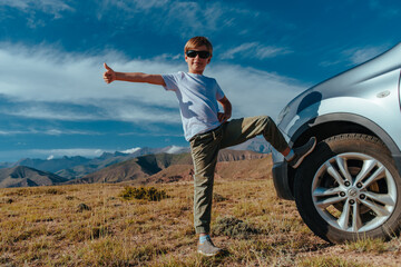 Canvas Print - Happy boy in sunglasses stands by the car in the mountains and gives a thumbs up, travel concept