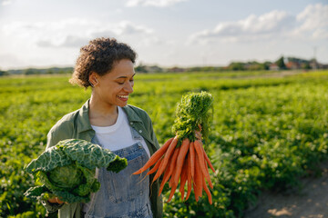 Wall Mural - Smiling female farmer standing with just harvested vegetables on the field background