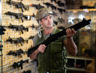 Wall Mural - European man in armor vest standing in salesroom of gun shop and selecting machine gun.