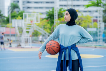 Wall Mural - Young asian muslim girl teen wearing hijab going to play basketball on the outdoor court in the morning with determination, Muslim sport concept.