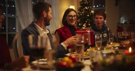 Wall Mural - Handsome Boyfriend Presenting a Festive Gift to His Beautiful Surprised Girlfriend. Family and Friends Gather at Home in the Evening for a Traditional Christmas Dinner with a Turkey Roast Feast