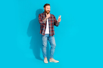 Wall Mural - Full size photo of minded thoughtful man with tattoo wear plaid shirt denim trousers look at smartphone isolated on blue color background