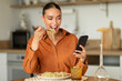 © Prostock-studio - Cheerful woman eating tasty homemade pasta and using cellphone while having dinner, lady scrolling social media feed
