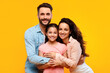 Happy european parents hugging their pretty daughter and smiling to camera on yellow background, studio shot