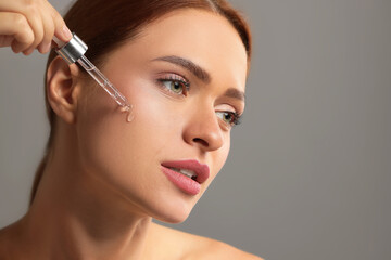 Poster - Beautiful young woman applying cosmetic serum onto her face on grey background, space for text