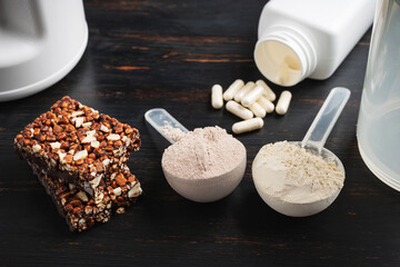 Two scoops of whey or soy protein powder, white capsules of amino acids, vitamins, creatine, protein chocolate bar, bodybuilding food supplements, sports nutrition on a dark wooden board