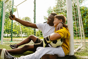 Wall Mural - African man taking selfie with his son while sitting on a football field