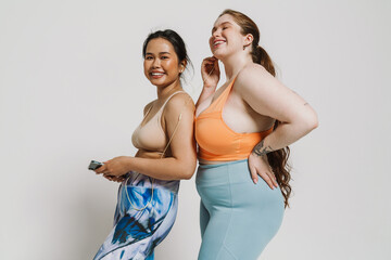 Wall Mural - Two smiling women listening music with earphones and mobile phone while standing isolated over white background