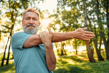 Wall Mural - Sportsman working out on nature. Portrait of charismatic athletic man of mature age doing arms exercises for warming up outdoors. Healthy man dressed in blue shirt confidently looking at camera.