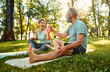 © HBS - Sport and recreation outdoors. Angle view of two family partners having rest and sitting on fitness mats in park. Smiling senior lady sharing bottle of cold fresh water with husband.