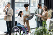 © LIGHTFIELD STUDIOS - cheerful interracial business people and disabled woman in wheelchair using devices in coworking