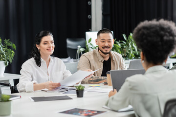 cheerful interracial business people looking at female colleague  while working in modern coworking