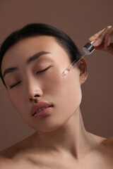 Canvas Print - Beautiful young woman applying cosmetic serum onto her face on brown background, closeup