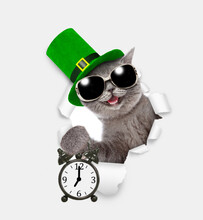 Saint Patricks Day Concept. Happy  Cat Wearing Sunglasses And Green Leprechauns Hat Looks Through The Hole In White Paper And Shows Alarm Clock