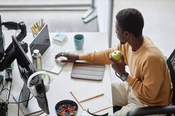 High angle view of programmer eating green apple and working on laptop in IT office