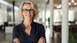 Eine selbstbewusste Geschäftsfrau mittleren Alters mit Brille steht lächelnd im Büro, die Arme verschränkt