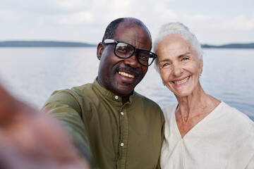 Selfie portrait of happy senior couple smiling at camera while standing outdoors against the lake