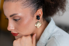 Crop Side View Of Young Thoughtful Female With Makeup Touching Chin While Resting And Listening To Music Over Wireless Ear Bud In Room Against Blurred Background