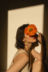 Wall Mural - Young girl hiding face with red poppy flower on pastel beige background with aesthetic sunlight shadow