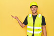 Latino worker in yellow vest and helmet showing a copy space on a palm and holding another hand on waist.